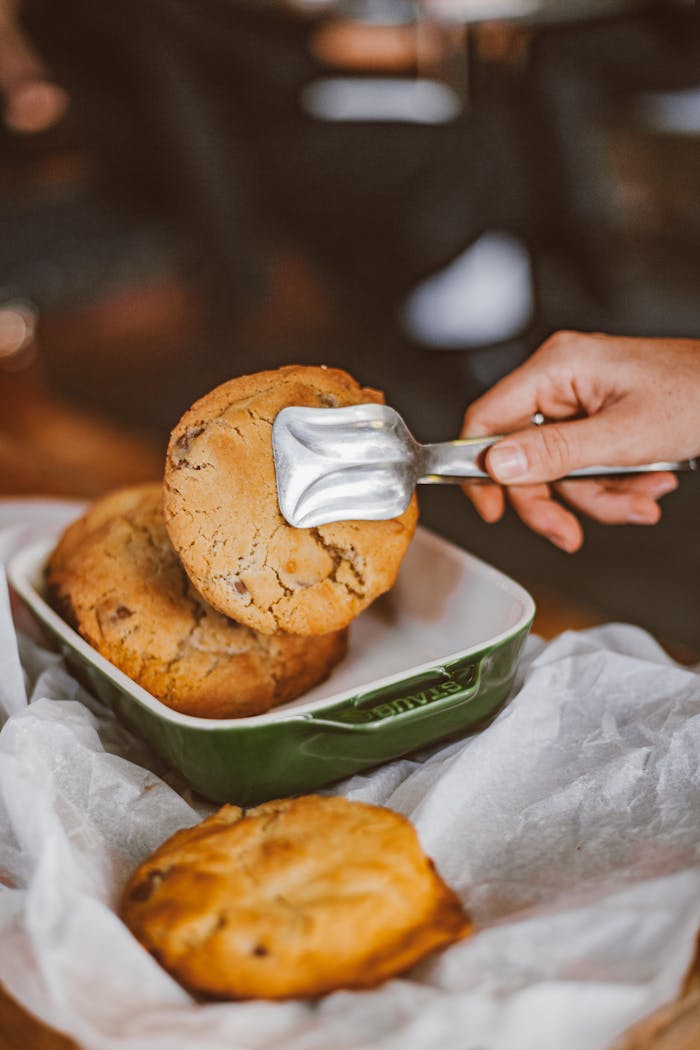 A hand using tongs to serve warm cookies on wax paper.